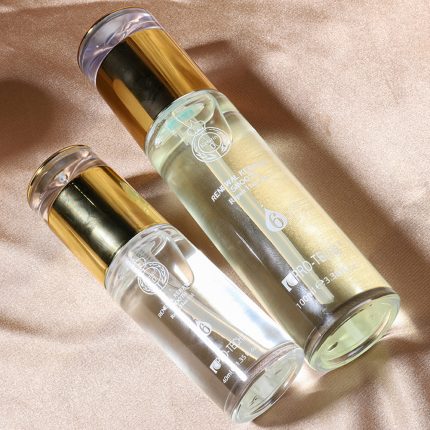 Two cylindrical glass bottles with gold caps lying on a beige fabric, containing clear and light‑yellow liquids and labeled product text visible on the bottles (cosmetic hair/skin serum).