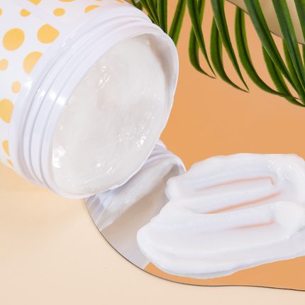 Open jar of white cream with lotion spread on a reflective surface; orange backdrop and green leaves in the background.