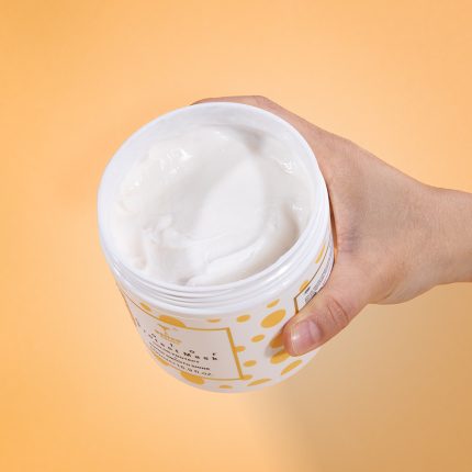 Hand holding an open jar of white moisturizer with a yellow-dotted label, against an orange gradient background.