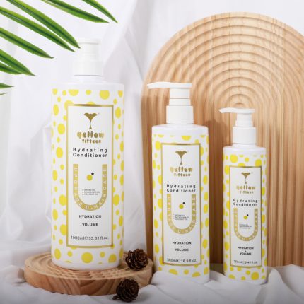 Three white pump bottles with yellow polka-dot labels reading Hydrating Conditioner, arranged on a round wooden display with a white fabric backdrop and green plant leaves nearby.