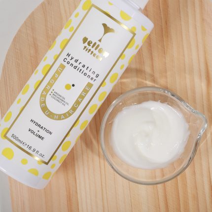 White bottle with yellow polka dots labeled Hydrating Conditioner beside a glass bowl of white cream on a wooden surface.
