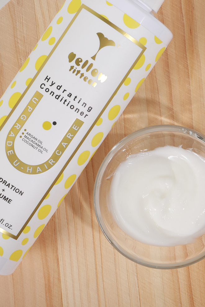White bottle with yellow polka dots labeled Hello Hydrating Conditioner, diagonally placed on a wooden surface beside a bowl of conditioner.