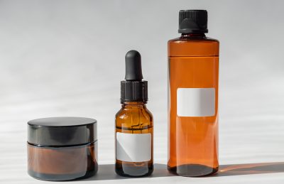 Amber glass skincare trio on a light surface: a small jar with a black lid, a dropper bottle, and a tall bottle with blank label(s).