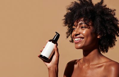 Joyful woman with natural curls holds a pump bottle of skincare product against a beige background.