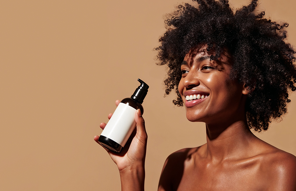 Joyful woman with natural curls holds a pump bottle of skincare product against a beige background.