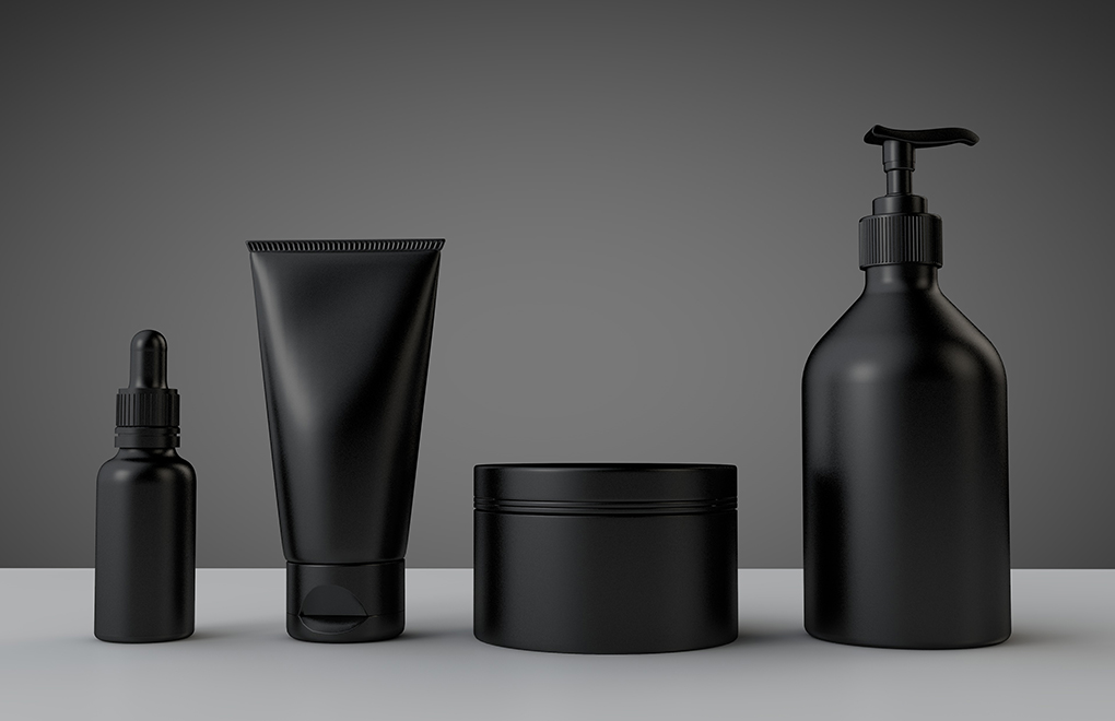 Set of matte black skincare containers on a gray surface: dropper bottle, squeezy tube, jar, and pump bottle in a row.