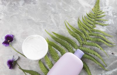 Lavender shampoo bottle and open cream jar on a gray surface with ferns and purple pansies.