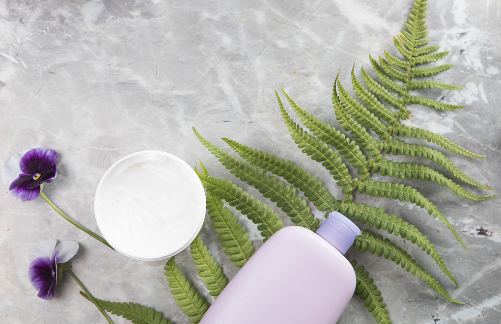 Lavender shampoo bottle and open cream jar on a gray surface with ferns and purple pansies.