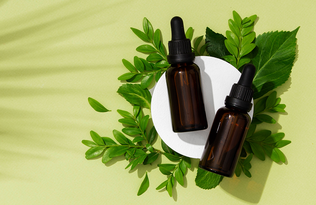 Two brown glass dropper bottles surrounded by green leaves on a white circular platform against a pale yellow background.