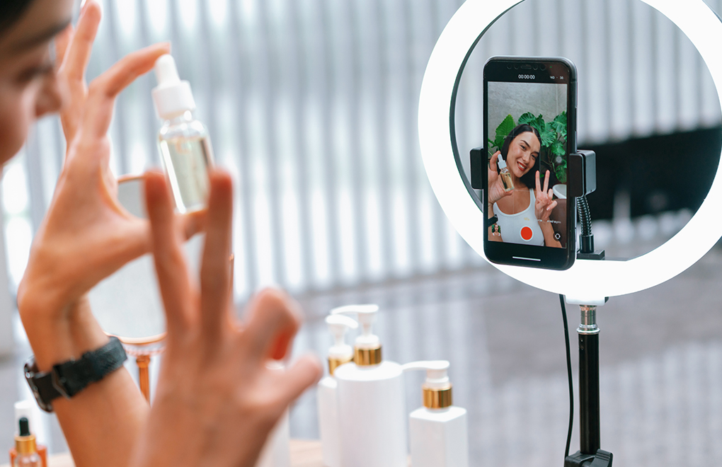 Beauty influencer filming a skincare/tutorial setup with a spray bottle; a smartphone on a stand faces the camera under a ring light showing the person smiling and making a peace signisy.