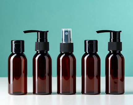 Five dark amber bottles with pump and spray tops arranged in a row on a white surface against a turquoise background.