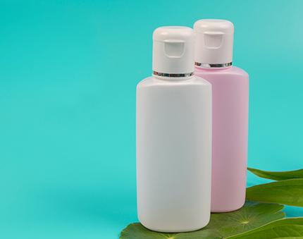 Two plastic lotion bottles in white and pink with flip-top caps on a turquoise background, with green leaves nearby.