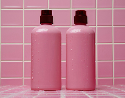 Two pink plastic bottles with dark caps stand side by side on a pink tiled surface and backdrop.