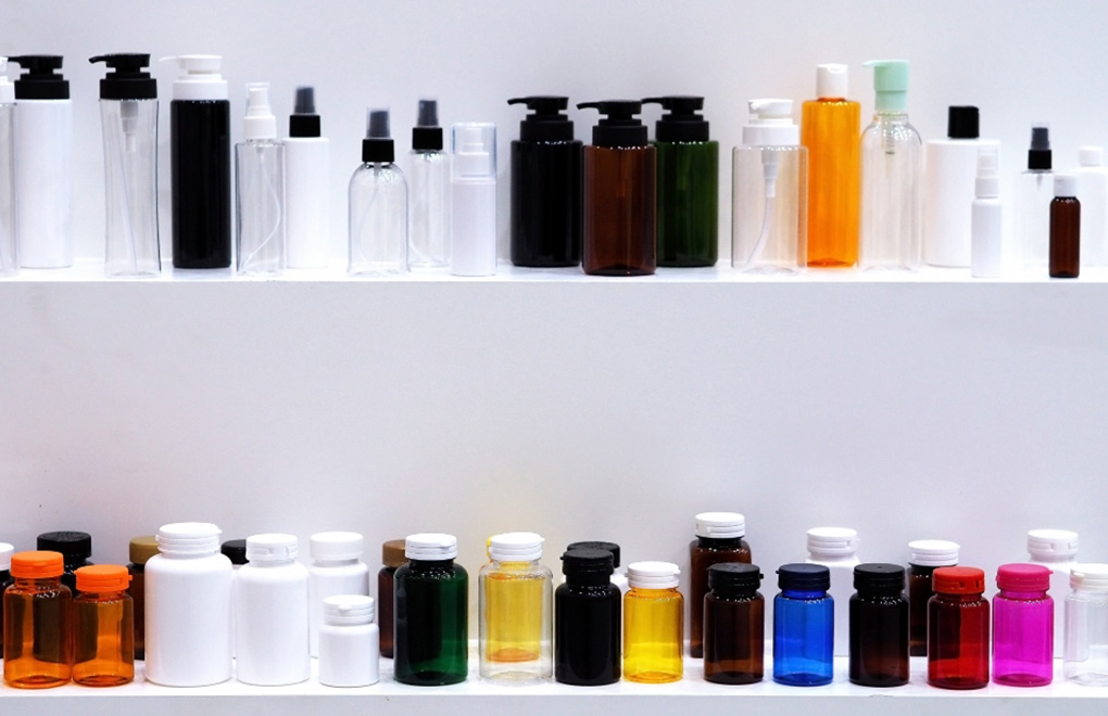 Row of assorted cosmetic and essential oil bottles in clear, amber, green, and colored containers on white shelves with pumps and caps.