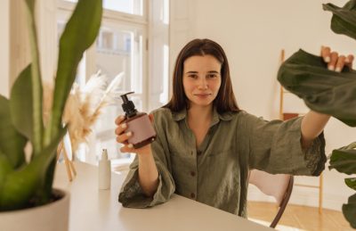 Woman sits at a bright desk, holding a pump bottle of lotion and reaching toward a large plant in a sunlit room.