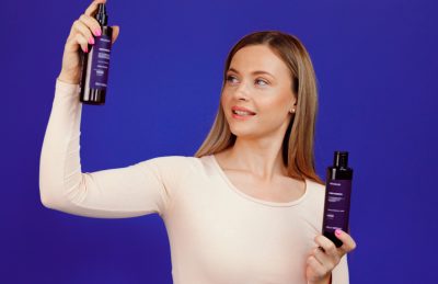 Woman in a light sweater holds up two dark purple haircare spray bottles against a blue backdrop, looking at the left bottle.