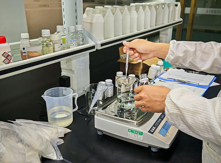 Lab technician uses a pipette to transfer liquid into a beaker on an electronic balance at a chemistry bench.