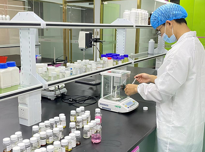 Lab technician in a white coat, hairnet, and mask weighs a sample on a digital balance at a lab bench with rows of chemical bottles nearby.