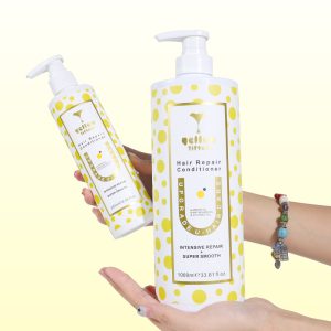 Large white bottle of Yellow Yitters Hair Repair Conditioner with yellow polka dots, held by a hand with bracelets; a smaller matching bottle appears in the background against a pale yellow backdrop.