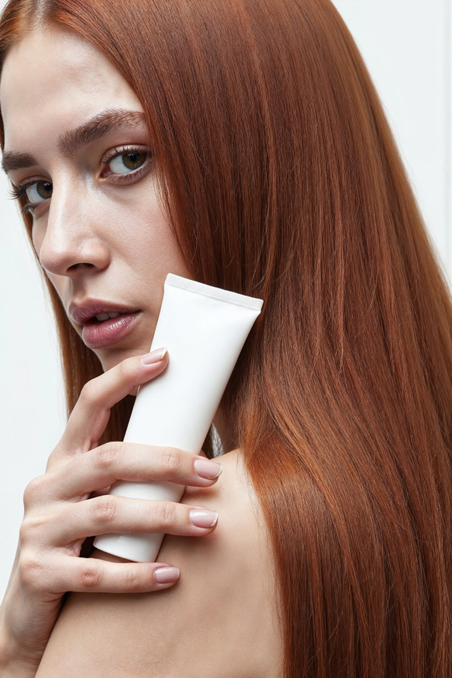 Woman with long red hair holding a white tube of cream near her face, as if applying skincare on her shoulder or neck area at close range.