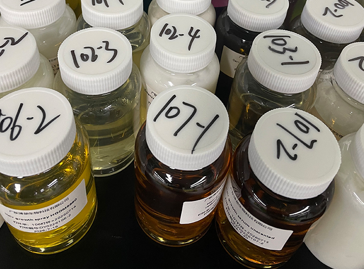 Row of small glass bottles with white screw caps and handwritten labels, containing yellow to amber liquids and arranged on a counter in a lab setting.